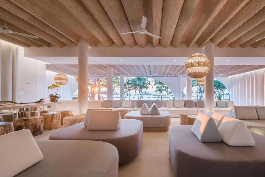 Open beach-resort lounge with timber ceiling, pale upholstered seating and a view toward palm trees and the sea