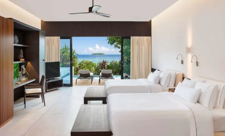 Resort guest room with two beds, a bench, desk, shelving and an ocean-facing opening